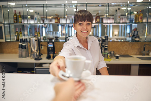 Cheeful pretty barmaid serving client with hot drink