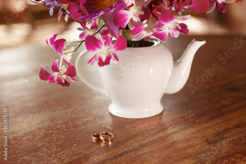 Wedding rings on woodn table near vase wth purple orchid flowers © Yevhenii