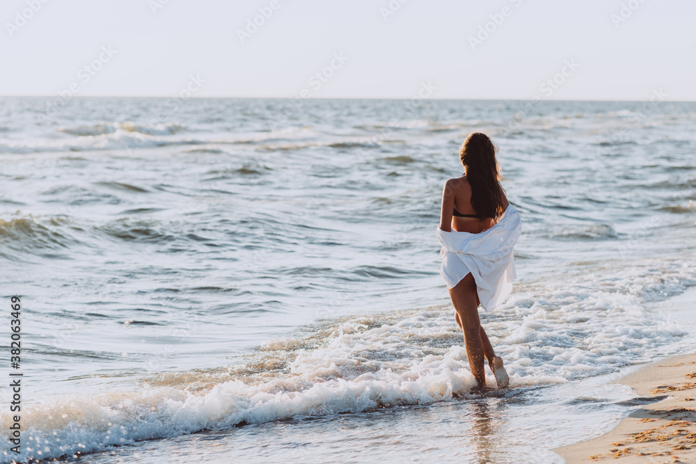 Young beautiful slender woman in a black bikini and white shirt on a tanned body on the beach in the waves, back view. Soft selective focus. Young beautiful slender woman in a black bikini and white shirt on a tanned body on the beach in the waves, back view. Soft selective focus.