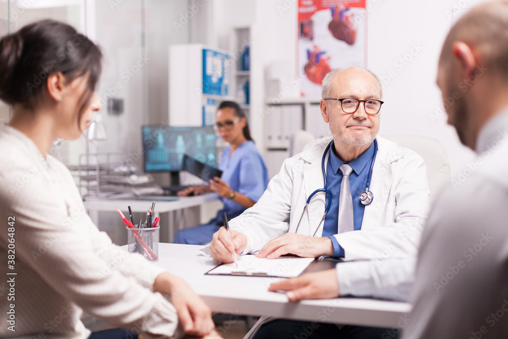 Senior medical practitioner taking notes on clipboard during consultation of married couple in hospital office. Doctor looking at male patient while wearing white coat and stethoscope. Senior medical practitioner taking notes on clipboard during consultation of married couple in hospital office. Doctor looking at male patient while wearing white coat and stethoscope.