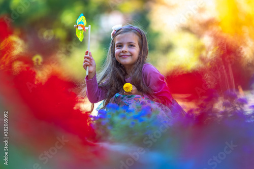 Happy young girl playing and blowing pinwheel in park with colorful flowers in slow motion