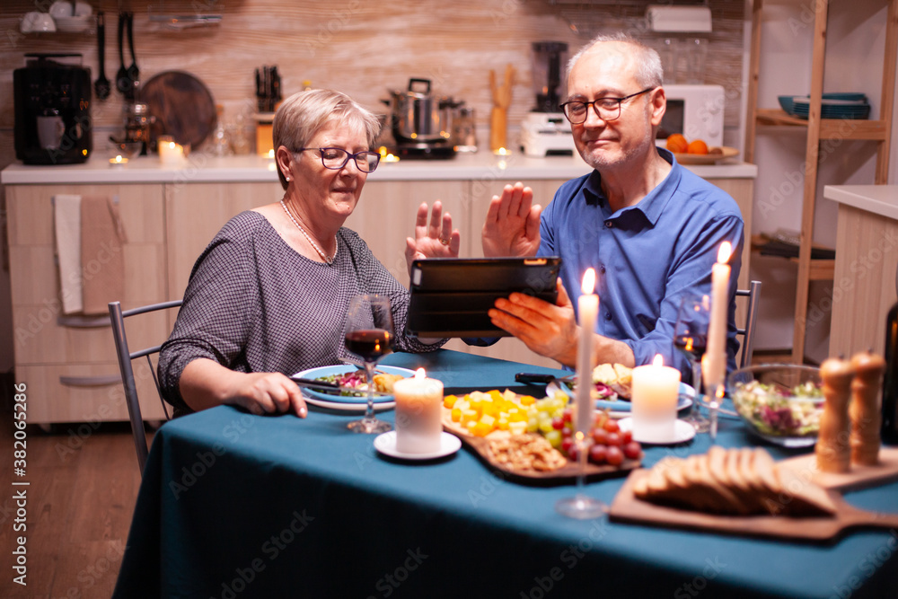 Smiling senior couple waving at camera of tablet pc during video call and dinner in kitchen. Couple sitting at the table, browsing, talking, using internet, celebrating their anniversary in dining
