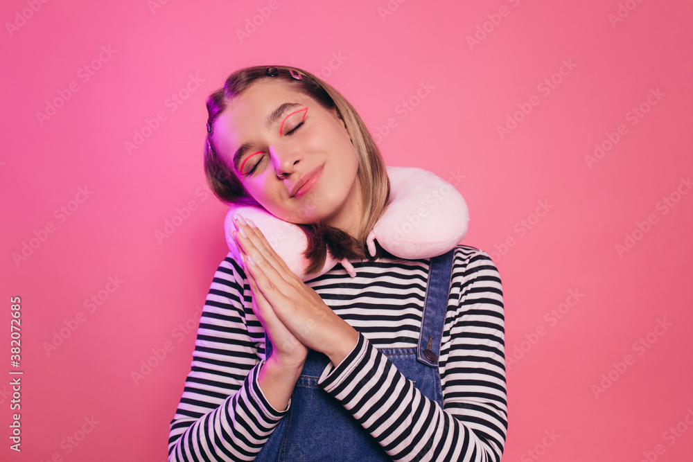 Attractive calm young woman stands isolated over pink background with closed eyes, keeps palms together, her head rests on travel pillow on her neck. Rest concept. Attractive calm young woman stands isolated over pink background with closed eyes, keeps palms together, her head rests on travel pillow on her neck. Rest concept.