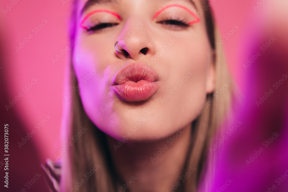 Attractive sexy young woman holding camera, blowing a kiss with closed eyes. Isolated over pink background. Lips in a tubule. Kissing concept. Pink cat eyes makeup. Attractive sexy young woman holding camera, blowing a kiss with closed eyes. Isolated over pink background. Lips in a tubule. Kissing concept. Pink cat eyes makeup.