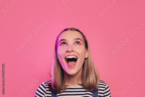 Happy extremely excited pretty young woman with open mouth looking up. Beautiful teenage girl saw something over her head. Isolated over pink studio background. Casual style.