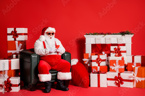 Portrait of his he nice attractive funky fat amazed Santa using device browsing web sitting in armchair isolated over bright vivid shine vibrant red color background