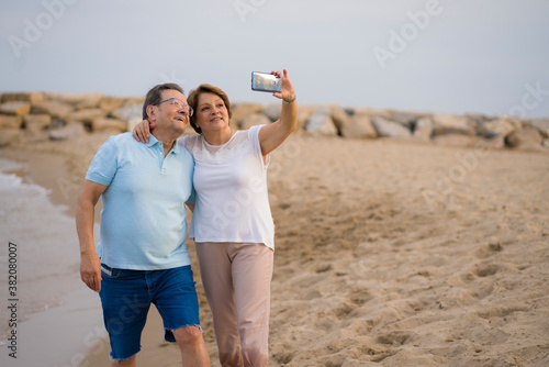 happy pensioner woman and her husband taking romantic walk taking selfie - happy retired mature couple walking on the beach during holidays taking self portrait with mobile phone photo