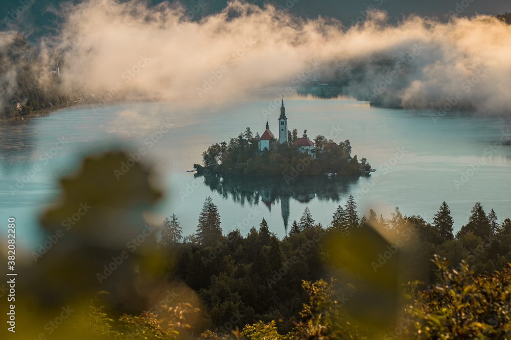 Panoramic photo of lake Bled island with church on a cold hazy foggy early autumn morning from Ojstrica vantage point. Visible leaves around the dreamy lake.