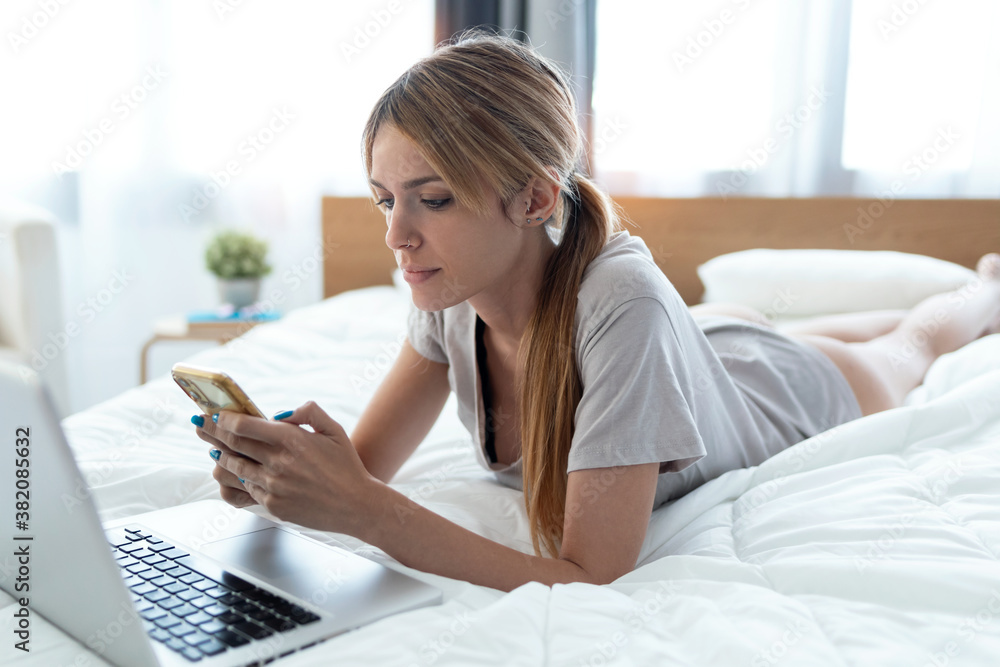 Beautiful young woman using her smart phone while working with laptop lying on the bed at home. Beautiful young woman using her smart phone while working with laptop lying on the bed at home.