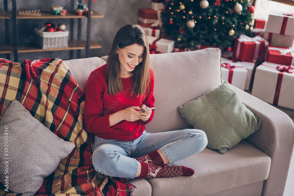 Fototapeta premium Full length photo of positive girl sit couch text cellphone wear red sweater in house indoors with x-mas christmas decoration