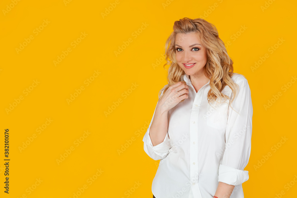 Blond woman in a white shirt, middle-aged European girl, neatly combs her hair with her hand, tilts her head and smiles sweetly at the camera with a coquettish caring look, yellow background. Blond woman in a white shirt, middle-aged European girl, neatly combs her hair with her hand, tilts her head and smiles sweetly at the camera with a coquettish caring look, yellow background.