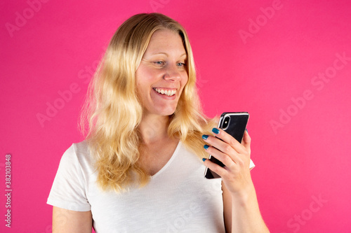 Blonde young woman holding smartphone while making a video call. Isolated on pink background. Looking happy.