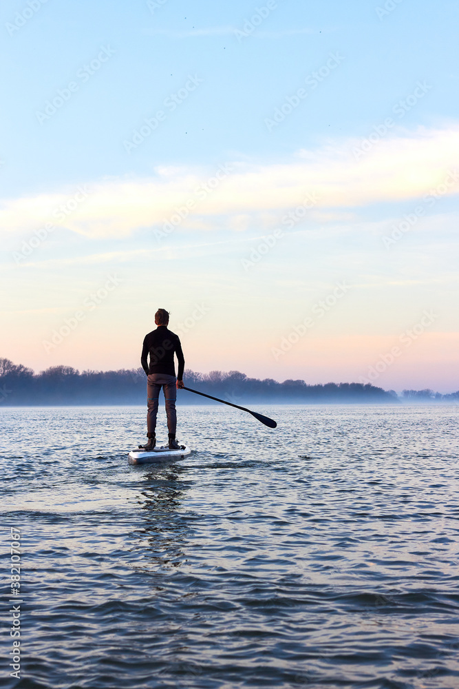 Back view on teenager boy rowing on SUP (stand up paddle board) in autumn Danube river at foggy morning against the backdrop of autumn trees without leaves