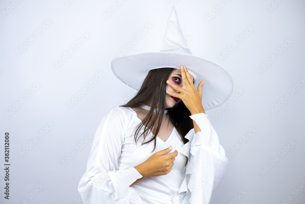 Woman wearing witch costume over isolated white background peeking in shock covering face and eyes with hand, looking through fingers with embarrassed expression Woman wearing witch costume over isolated white background peeking in shock covering face and eyes with hand, looking through fingers with embarrassed expression