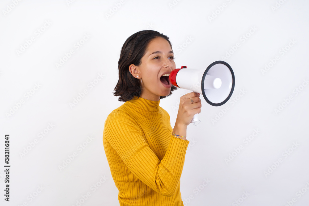 Young hispanic girl with short hair wearing casual yellow sweater isolated over white background Through Megaphone with Available Copy Space Young hispanic girl with short hair wearing casual yellow sweater isolated over white background Through Megaphone with Available Copy Space
