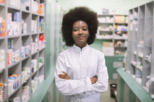 Smiling African American woman pharmacist or chemist posing to camera with arms crossed, while standing in interior of modern light pharmacy. Pharmacy and medicine concept.