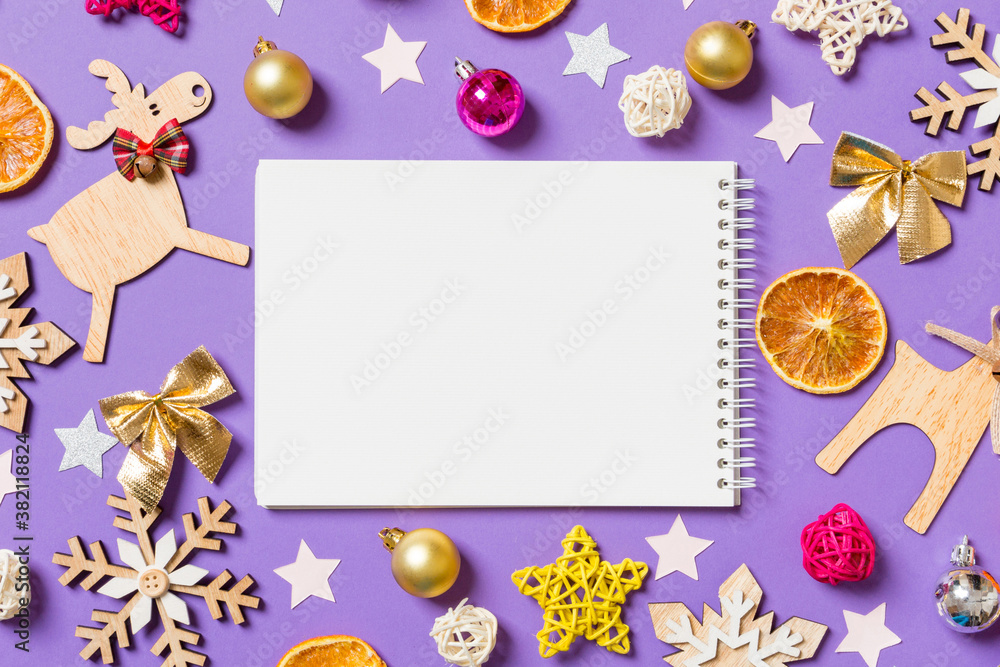Fototapeta premium Top view of notebook surrounded with New Year toys and decorations on purple background. Christmas time concept
