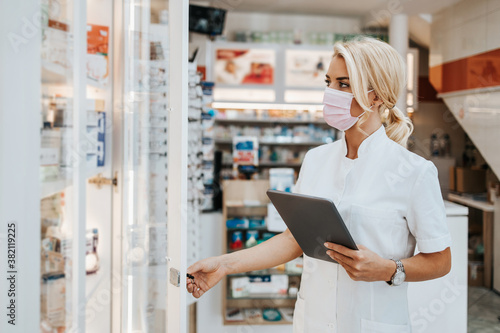 Young and attractive female pharmacist with face protective mask working in drugstore. She is confident and serious. Covid-19 open for business concept.