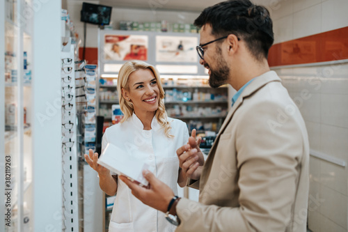 Young business man choosing and buying drugs in a drugstore while talking with attractive female pharmacist. She helping him with expert advices.