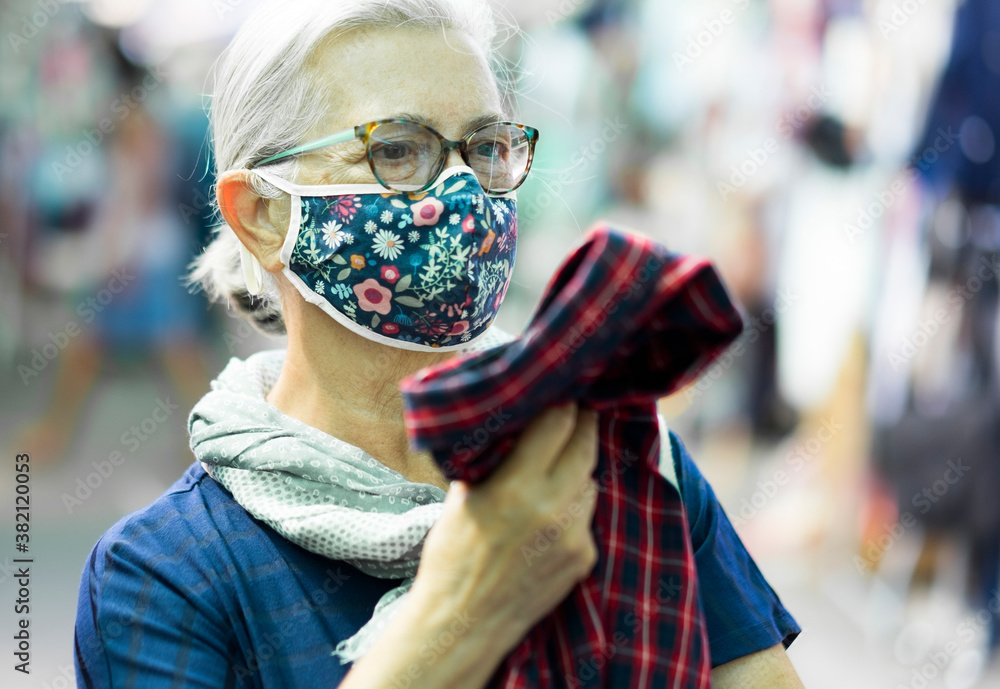 Portrait of an elderly woman wearing a medical mask due to coronavirus while shopping for a used shirt at the flea market.