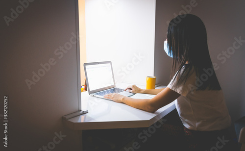 Side view of girl with black hair in white shirt and medical mask on sitting at home and typing on keyboard. Study at home concept photo