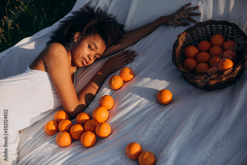 Dark-skinned model lies on white cloth around lot of oranges . Afro-American woman with natural makeup is holding an orange on a white cloth