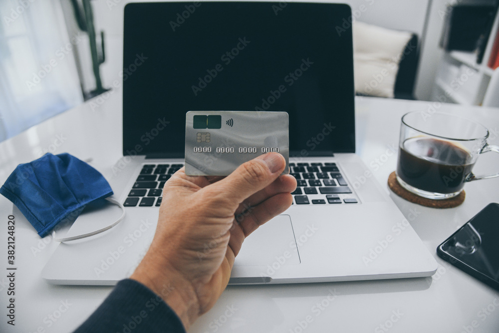Male hand with credit card. Home workstation during covid-19 lockdown. Home shopping due to virus pandemic emergency. Protective mask on laptop. Smart working, prevention health concept. Interior view Male hand with credit card. Home workstation during covid-19 lockdown. Home shopping due to virus pandemic emergency. Protective mask on laptop. Smart working, prevention health concept. Interior view
