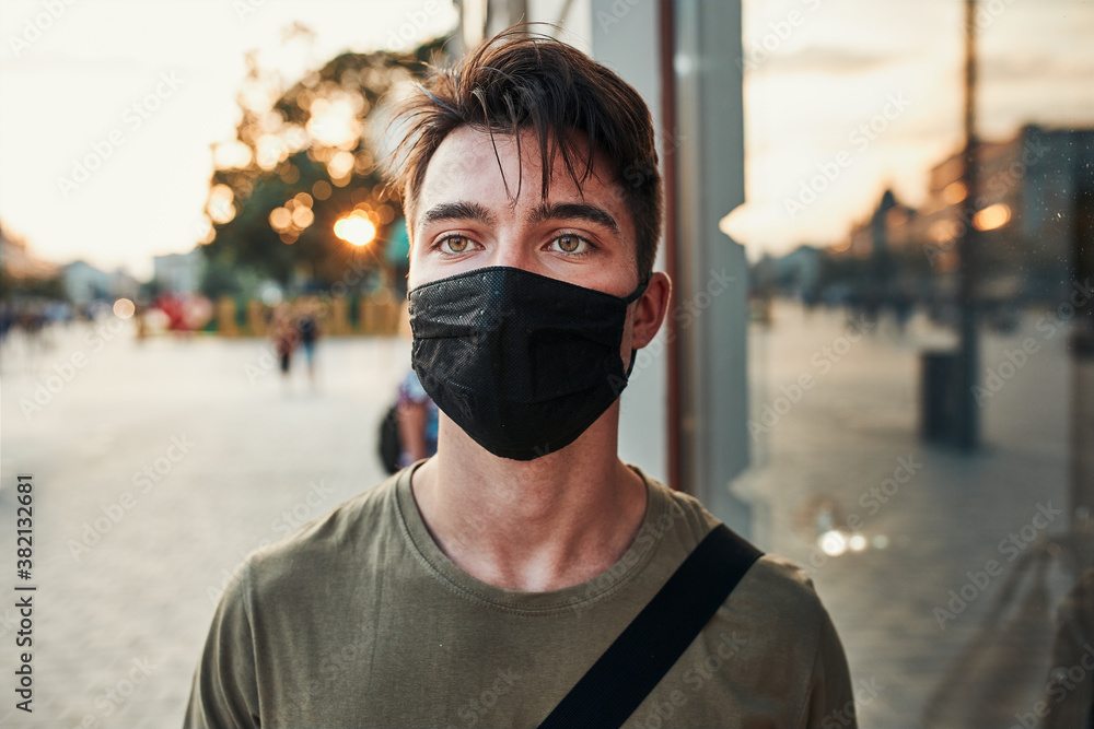 Young man walking along a store front in the city center in the evening wearing the face mask to avoid virus infection and to prevent the spread of disease in time of coronavirus Young man walking along a store front in the city center in the evening wearing the face mask to avoid virus infection and to prevent the spread of disease in time of coronavirus