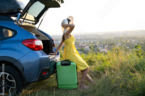 Young woman resting on a green suitcase near her car in summer nature. Travel and vacations concept.