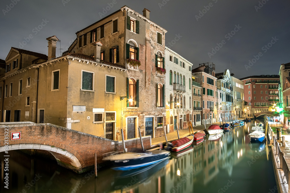 Evening view of illuminated old buildings, bridges, floating boats and light reflections in canals water in Venice, Italy.