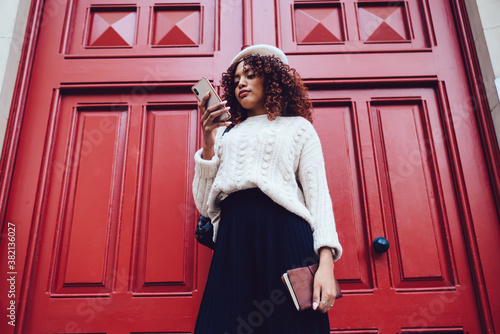 Beautiful curly hipster girl millennial sending mails and chatting via application on mobile phone on urban settings, positive dark skinned woman influencer searching and share content on smartphone © BullRun