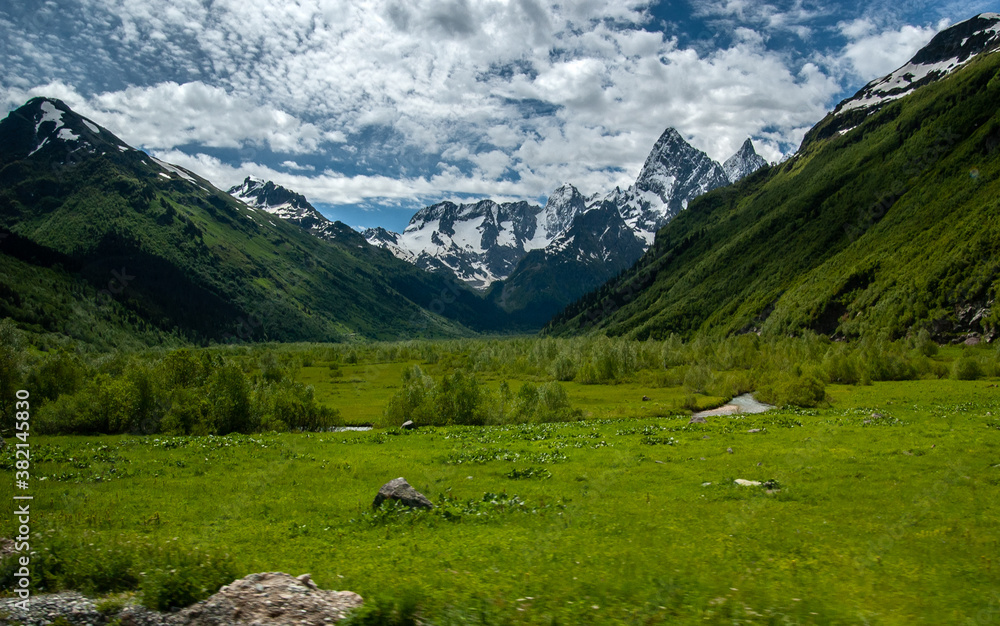 Valley with low birches. Creek among Emerald grass. Green mountains with forest and rocky snowy mountains surround the valley, blue sky. Caucasus. Russia. Valley with low birches. Creek among Emerald grass. Green mountains with forest and rocky snowy mountains surround the valley, blue sky. Caucasus. Russia.