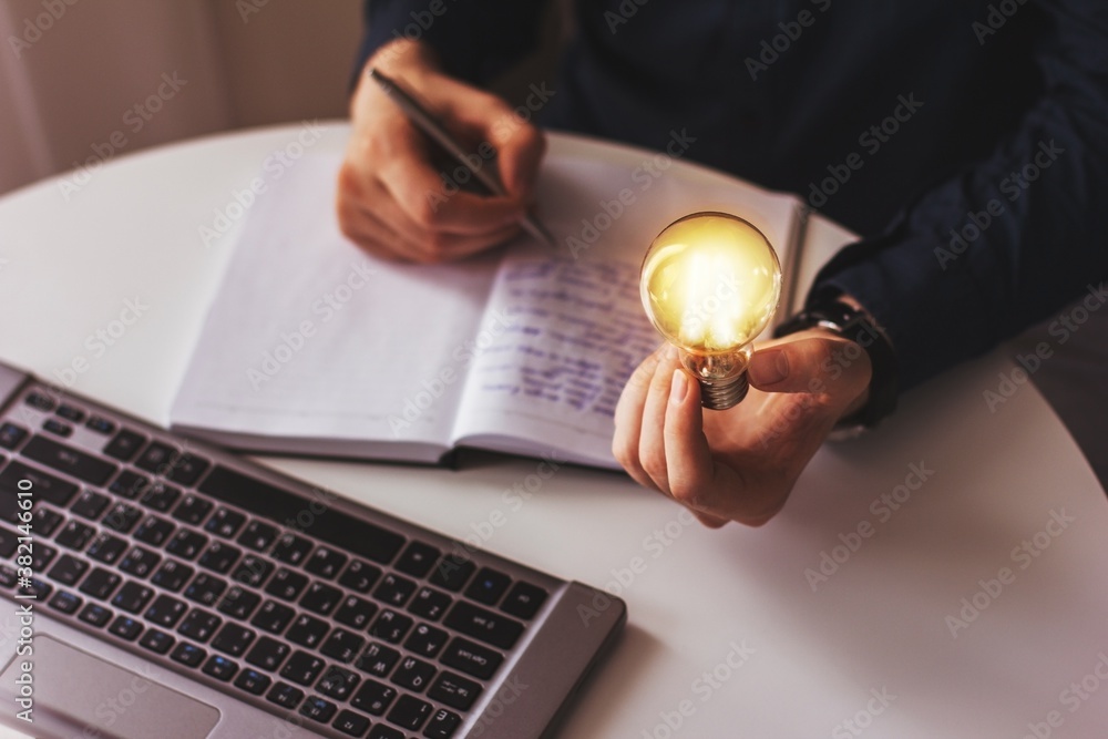 The hand of a business man holding a lighted light bulb, and writing in a notebook. Idea, innovation and inspiration with a light bulb that shines with brilliance