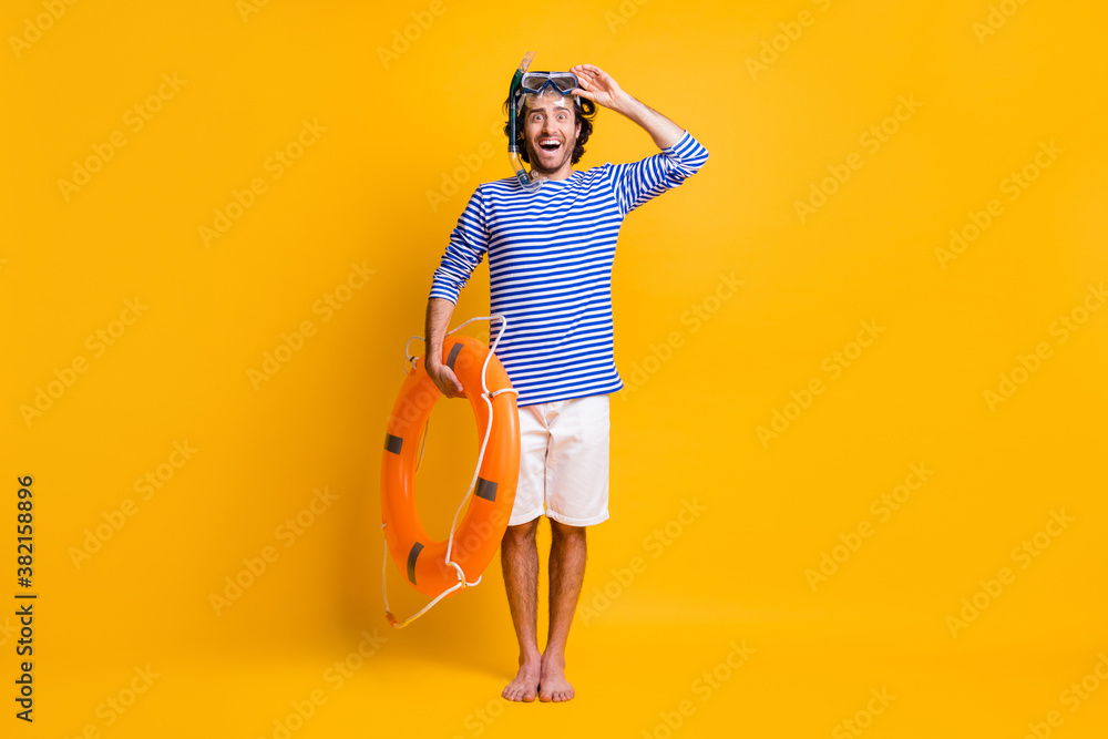 Ful size photo of excited guy hold life buoy touch scuba goggles wear blue striped shirt shorts isolated on yellow color background Ful size photo of excited guy hold life buoy touch scuba goggles wear blue striped shirt shorts isolated on yellow color background