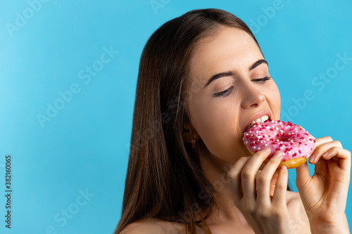 Close up portrait of a pleased pretty girl eating pink donuts isolated over blue background