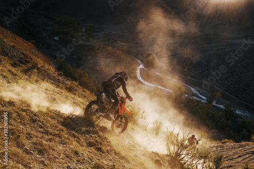 Extreme Rider climbing sand mountain top on off-road cross enduro motorcycle. Beautiful mountains landscape down on background, colourful autumn forest and river in sunshine