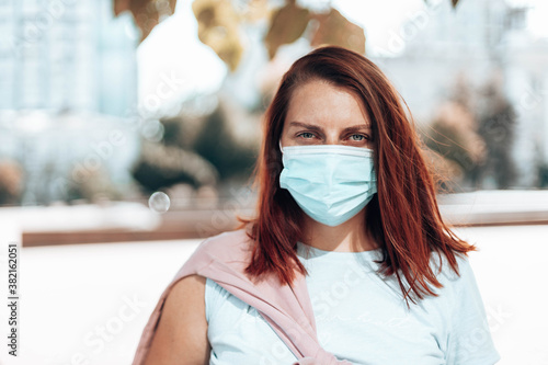 Portrait of a young beautiful red-haired girl in medical protective mask looking at the camera in park during the Coronavirus COVID-19 virus pandemic