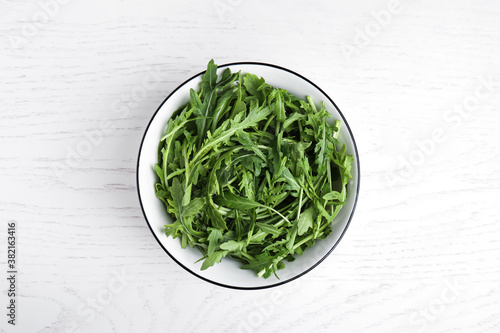 Fresh arugula in bowl on white wooden table, top view