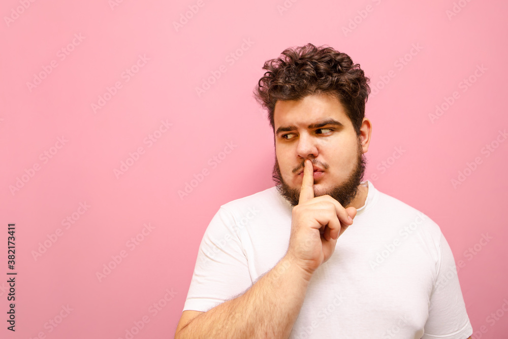 Serious guy in a white T-shirt and overweight looks aside with a pensive face and shows a sign of silence. Concentrated fat man with a beard shows the gesture Serious guy in a white T-shirt and overweight looks aside with a pensive face and shows a sign of silence. Concentrated fat man with a beard shows the gesture