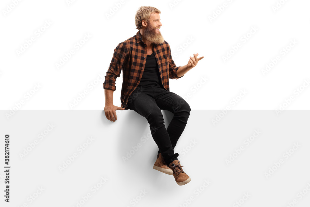 Young bearded man sitting on a white panel board and gesturing with hand