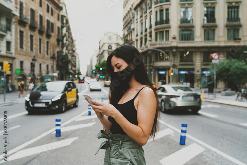 Young caucasian female in mask using online map application on a mobile phone to make a route. Woman wearing mask answering email by a mobile phone while standing on a road street background.