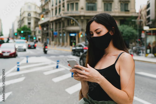 Young stylish woman in a black mask ordering a taxi by a mobile app on a smartphone to go home avoiding public transport. Woman wearing protective mask while standing on a street with a mobile phone.