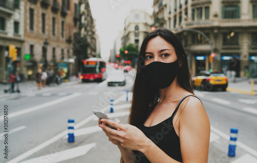 Portrait of a brunette woman wearing mask waiting for a taxi she ordered by the mobile phone application. Young woman in a mask looking aside while standing on a crossroad with a mobile phone in hands © JKstock