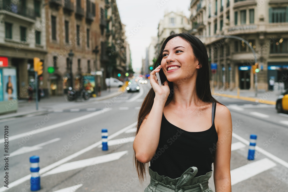 Cheerful caucasian hipster girl standing at the city street and talking on her smartphone. Young smiling woman with long dark hair in trendy outfit having phone conversation while looking aside