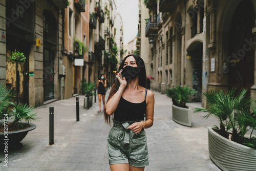 Happy young woman enjoying the street walking after the quarantine period in the city. Attractive female wearing protective mask having mobile phone conversation while going down the street.