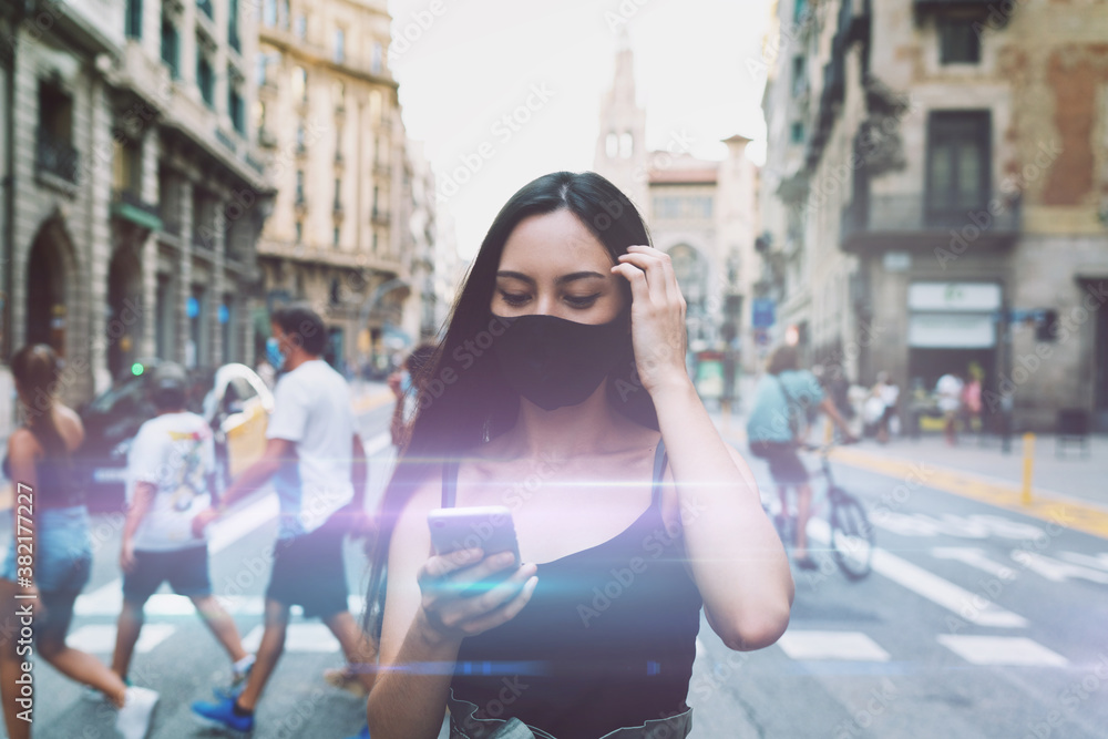 Attractive brunette female wearing medical mask using map app on mobile phone to find cafe to meet with friends. Young hipster girl in a protective mask checking messages on a smartphone. Flare light
