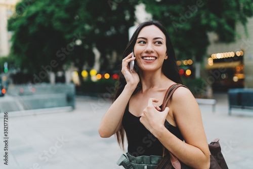 Young brunette caucasian woman with long hair is talking by a smartphone while walking down the street on a blurred urban background. Stylish student girl is chatting with friends by a mobile phone.