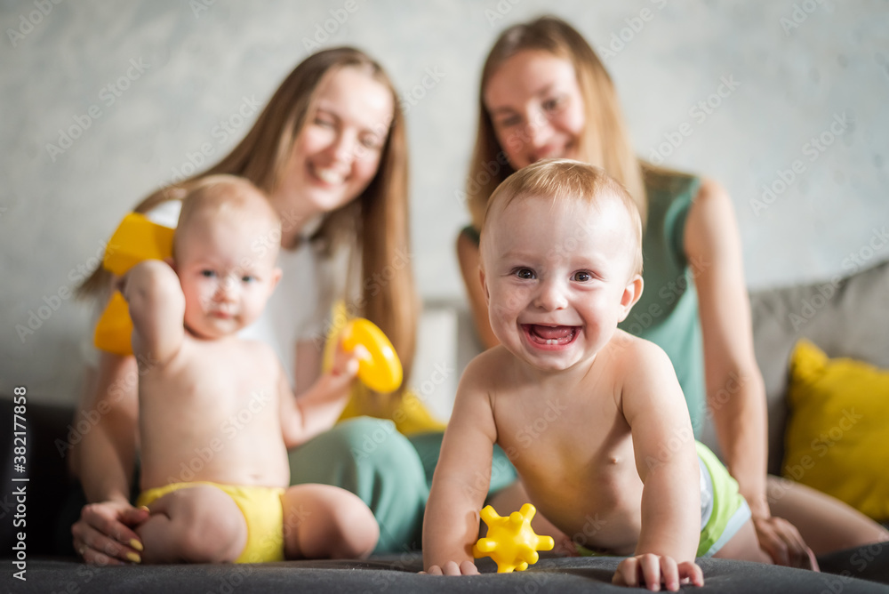 Two young mothers are playing with their happy kids in the room on the sofa. Happy baby laughs Two young mothers are playing with their happy kids in the room on the sofa. Happy baby laughs