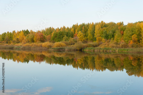 Autumnal lake shore with forest under blue sky. Colorful fall foliage reflecting on surface of calm water.