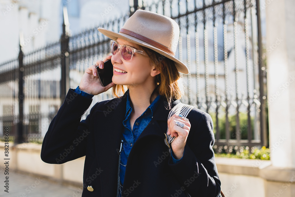 Fototapeta premium young stylish pretty woman smiling, happy, surprised, talking on her phone, dressed in dark blue coat and denim shirt, pink sunglasses, with a hat and backpack, sunny day, good weather, city street